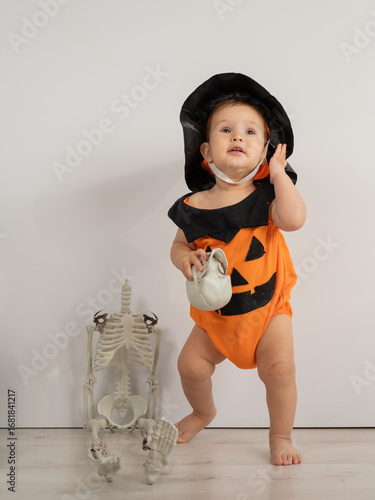 Cute caucasian boy in pumpkin costume for halloween holding skull on white background. 