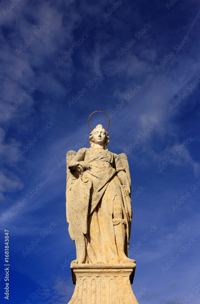 Fototapeta premium Historic statue of the Archangel Raphael, patron saint of Cordoba. It is located on the Roman Bridge which spans the Gaudalquivir River. Backed by Cirrus clouds on a deep blue sky. Copy space.