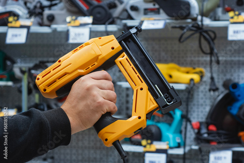Close-up of a person holding a yellow electric nail gun in a hardware store. Power tool for construction, woodworking, furniture assembly, and home renovation projects.