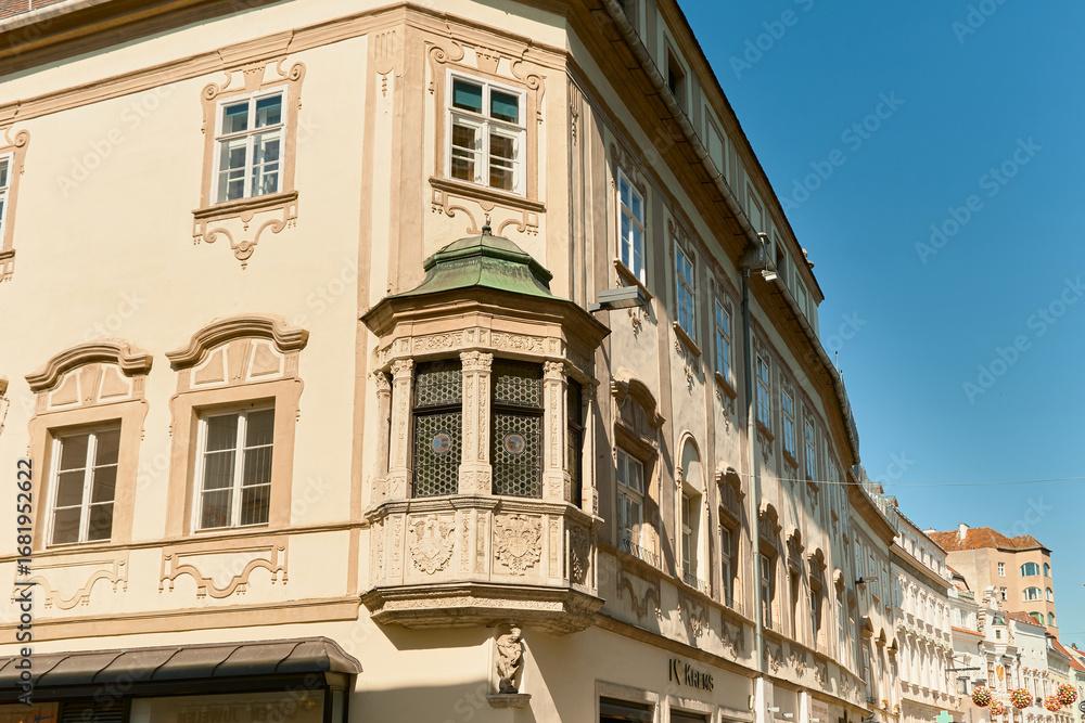Fototapeta premium Photo of a historic house in Wachau, Austria with green bay window, decorative facade, and a street lined with houses extending into the distance under a blue sky.