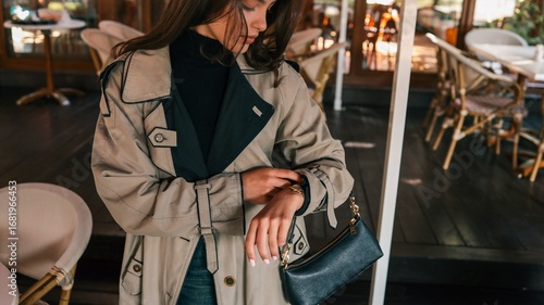 Tableau sur toile Young woman in beige trench coat looking at her wristwatch while standing outsid