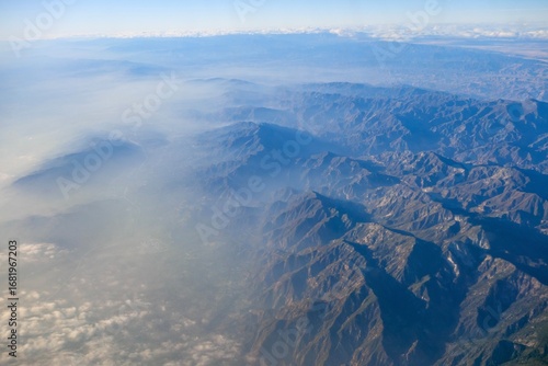 An aerial shot capturing the rugged beauty of the Angeles and San Gabriel Mountains with a hazy urban landscape visible below
