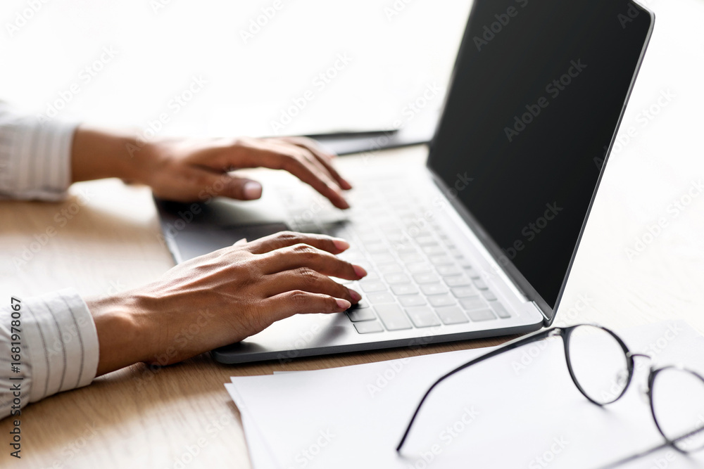 Fototapeta premium Modern work, remote office, business, devices for social distancing, covid-19 quarantine. Millennial african american woman typing on laptop with blank screen on table with glasses, cropped, close up