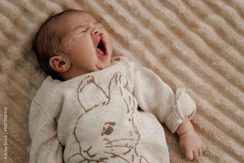 Bild auf Leinwand Yawning Baby in Rabbit Sweater on Beige Blanket
