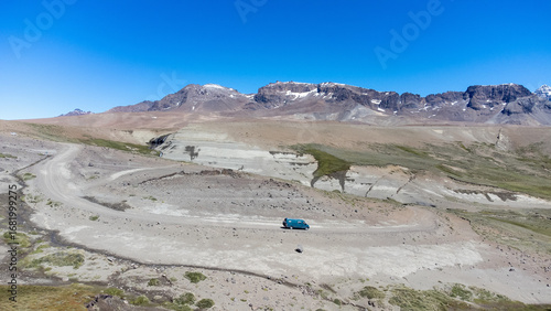 Drone view of a campervan driving on a dirt road in a desert mountain landscape with rocky cliffs and snow peaks. Concept of road trip, adventure and freedom travel