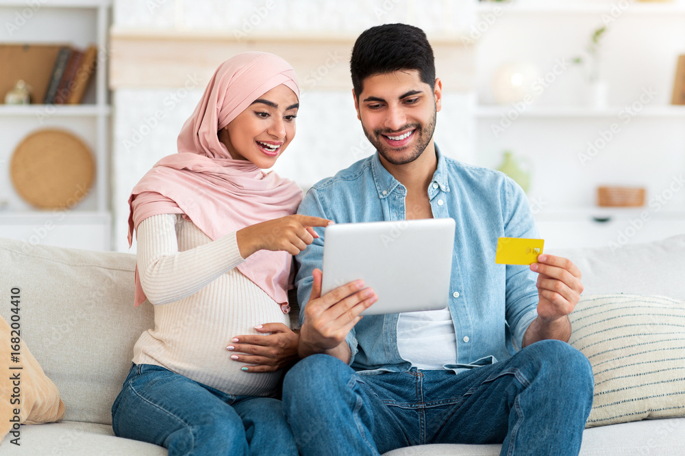 Fototapeta premium Excited pregnant islamic spouses shopping online with tablet and credit card at home while relaxing together on sofa in living room. Happy expectant arab couple buying baby clothes in internet