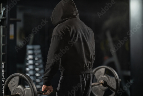A man wearing a hooded sweatshirt prepares to lift heavy weights in a gym. The atmosphere is focused and intense as he concentrates on his deadlift technique during a workout session.