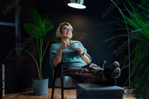 Middle aged woman relaxing with cup of hot drink in scandy style hygge interior home. Cozy comfortable winter or autumn weekends. Take a break and relax. Lady enjoy calm mood, well being alone.