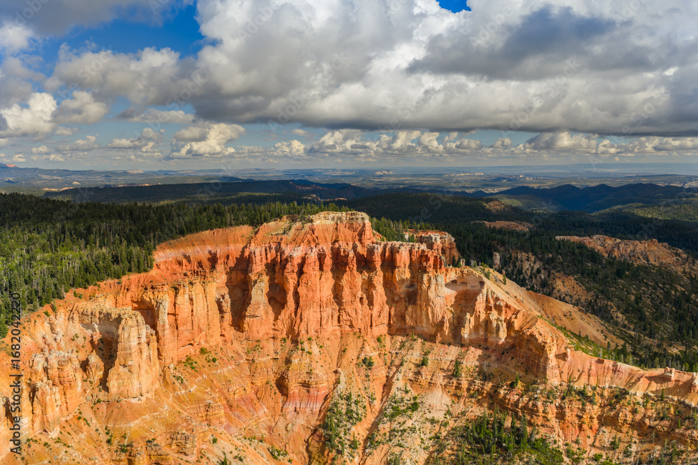 Fototapeta premium Dramatic red rock cliffs and hoodoos at Cedar Breaks National Monument in Utah, with forested mountains and clouds under a bright blue sky.