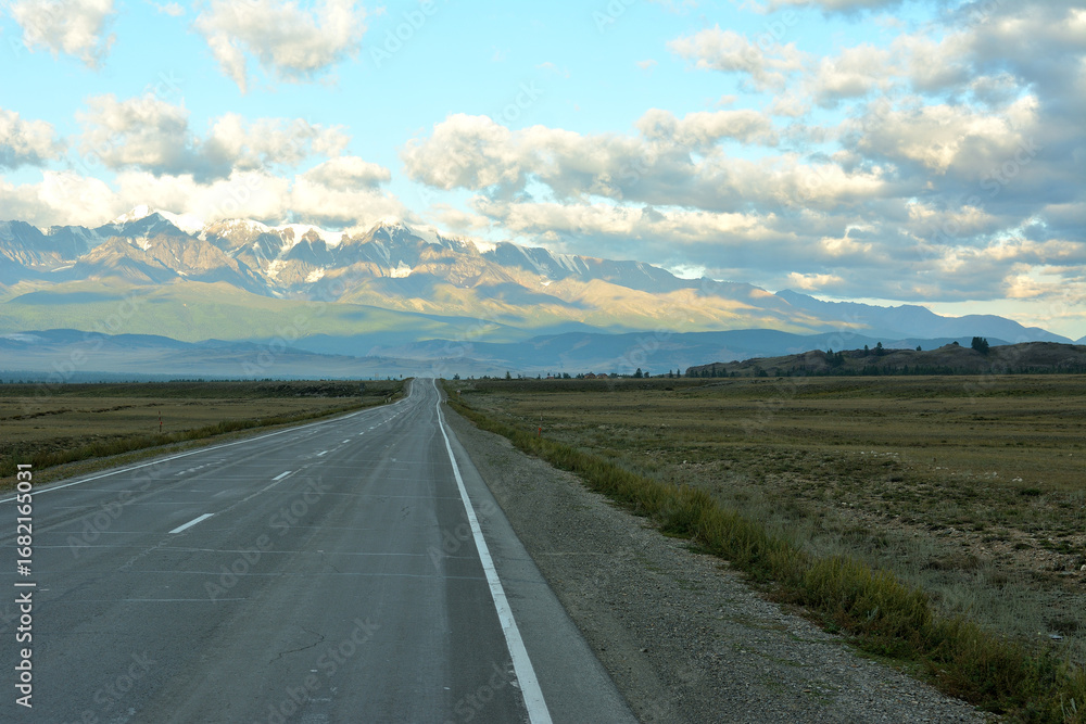 Ταπετσαρία A straight two-lane asphalt road leads up to high snow-capped mountains covered with clouds on a sunny summer morning