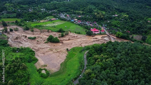 Drone aerial view of muddy floodwaters cutting through farmland and reaching a rural village, showing the dramatic contrast between lush green fields and disaster destruction.
