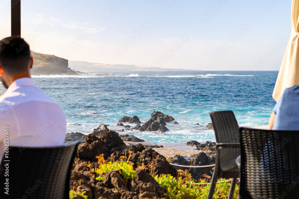 Obraz premium Tourists enjoy sea and mountain views along the volcanic coastline from an outdoor seaside cafe restaurant at Garachico, Spain, on the Canary Island of Tenerife.