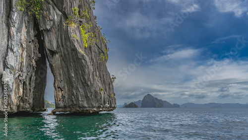 A picturesque cliff above the ocean. The gap between the steep slopes. Green vegetation on the rock. Mountains on the horizon. Blue sky, clouds. Philippines. Palawan. El Nido.