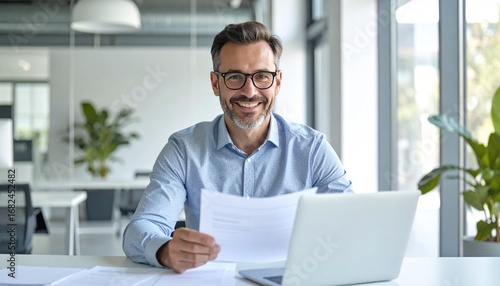 A smiling man with glasses sits at a desk with a laptop and papers in a bright office, gazing at the camera with a positive expression