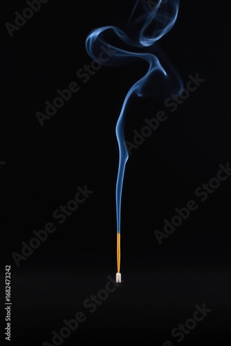 A single incense stick burning, emitting a wispy blue smoke trail against a stark black background