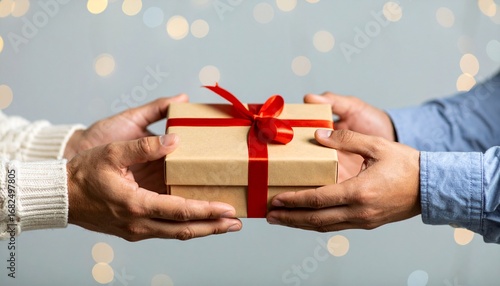 Two people's hands exchanging a beautifully wrapped gift box with a red ribbon against a festive blurred lights background, symbolizing generosity and celebration.