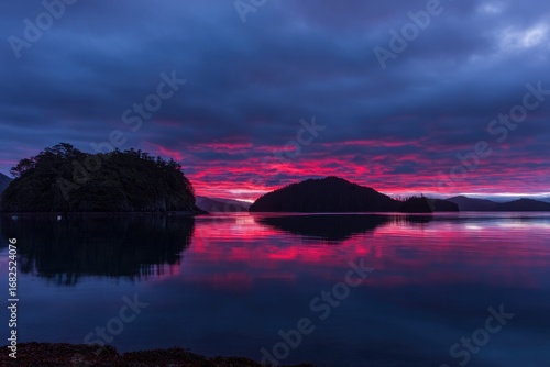 Vibrant sunrise reflected in calm water, dark islands silhouetted against fiery sky