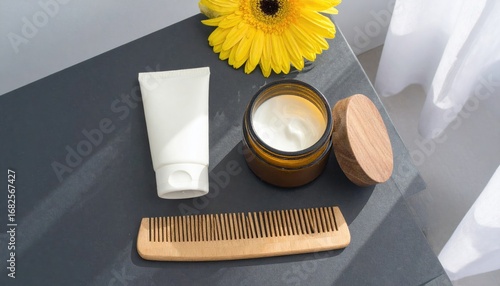 Cosmetic set with white tube, jar with wood lid, comb, and yellow flower on a grey background in natural light near a sheer white curtain