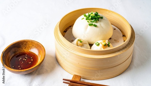 Steamed buns with greens in a bamboo container, accompanied by a small bowl of sauce and chopsticks on a white tablecloth