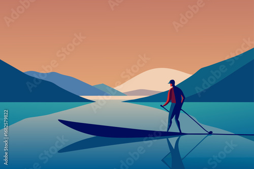 Man standing on a paddleboard with a paddle in a calm lake with mountains in the background during sunset