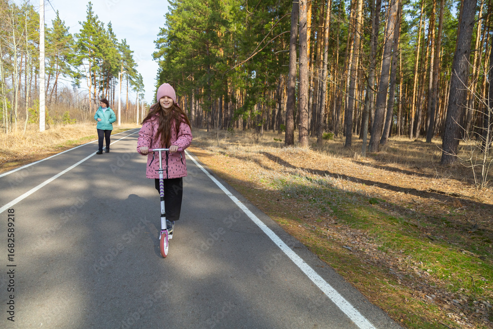 Fototapeta premium Little girl riding scooter while walking with her grandmother in the forest on a sunny day