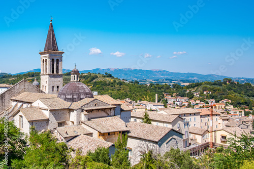 Spoleto Cathedral rises majestically amidst a picturesque cityscape, with its bell tower reaching into the clear blue sky and rolling green hills of Umbria, Italy
