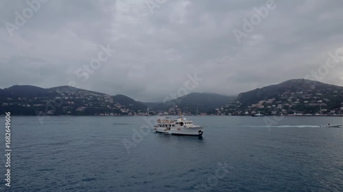 A tranquil drone capture of a vessel gently gliding on the Mediterranean off the coast of Mandelieu La Napoule, framed by lush hills and a cloudy sky, embodying the serene allure of the French Riviera