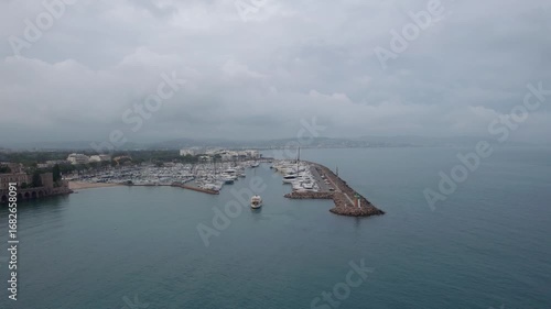 Drone footage showcases a marina in Mandelieu La Napoule, South of France, yachts docked along the vibrant pier, serene waters, coastal architecture, and hazy mountains under soft cloudy skies