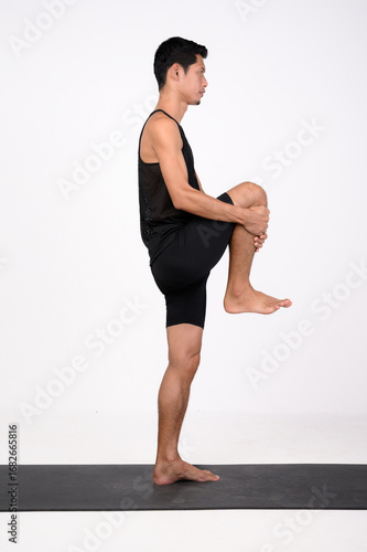 A male runner in black athletic wear performs a standing knee-to-chest stretch on a mat. A man wearing a black running suit is doing a dynamic leg stretch pose.