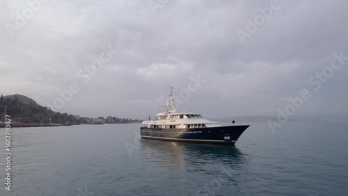 Drone footage reveals a luxury yacht cruising along the calm waters of the South of France, near a picturesque marina. The scene captures the elegance and serenity of maritime travel on a cloudy day