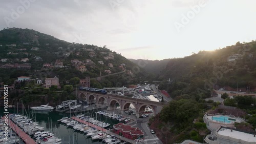 Drone footage captures a marina in Mandelieu La Napoule, South of France at sunset, featuring yachts, lush hills, and charming village architecture. setting creates a serene Mediterranean atmosphere