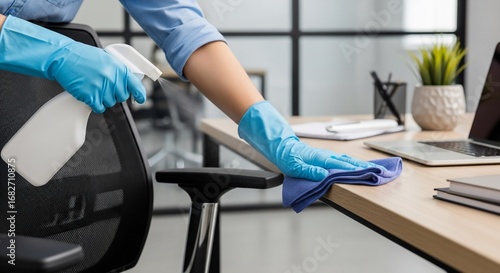 Person in blue gloves cleaning an office desk with a spray bottle and cloth, ensuring a hygienic workspace.