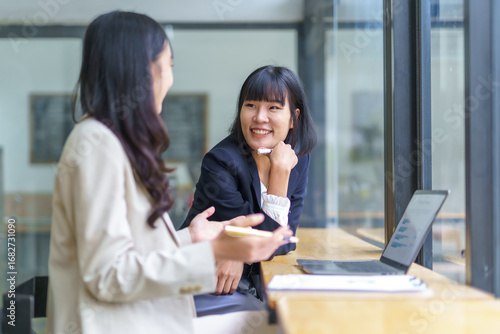 Two Asian businesswomen engaging in a discussion about marketing strategy while using a laptop in a modern office setting