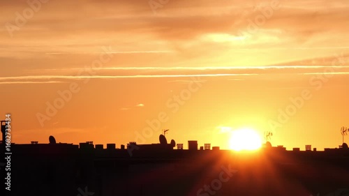A vibrant sunset over a city skyline. The sun is low on the horizon, casting warm orange and yellow hues across the sky. Silhouettes of buildings are visible.