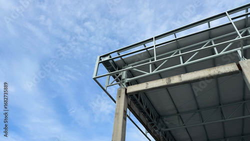 Corner view of concrete column and steel truss roof structure under construction with clear blue sky background, modern architecture and civil engineering detail.