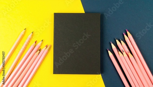 A black notebook rests between groups of pink pencils, set against a contrasting yellow and dark blue background, offering a clean, minimalist aesthetic