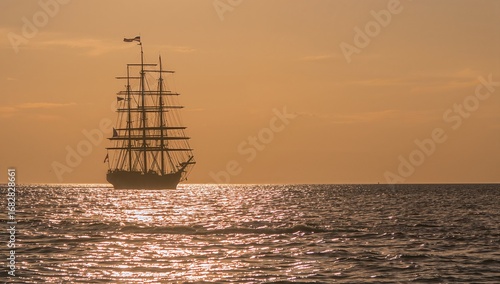 Gliding three-masted sailing vessel across calm sea at sunset, showing rigging and flag on mainmast