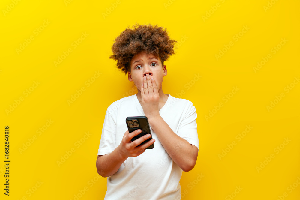 shocked african american boy in white t-shirt using smartphone and wondering on yellow isolated background, curly schoolboy choosing online and holding mobile phone in amazement shocked african american boy in white t-shirt using smartphone and wondering on yellow isolated background, curly schoolboy choosing online and holding mobile phone in amazement