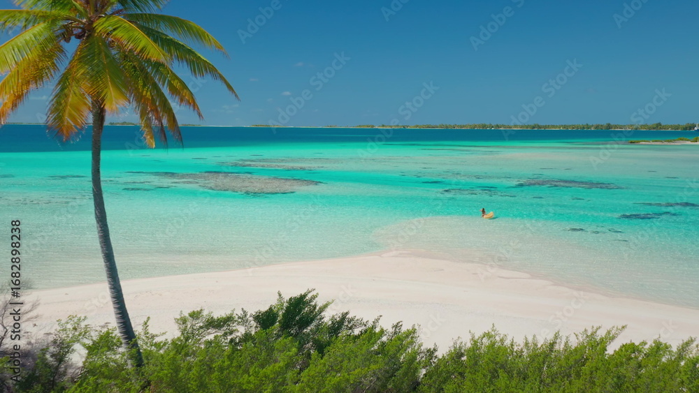 Fototapeta premium Woman happily floats on a yellow raft in crystal clear turquoise waters near a tropical island. With a palm tree and white sand beach in the background