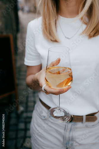 Young blonde woman enjoying glass of white wine while standing outdoors on charming cobbled street, dressed in casual white shirt and light jeans, radiating relaxed summer vibe. Glass of wine in hands