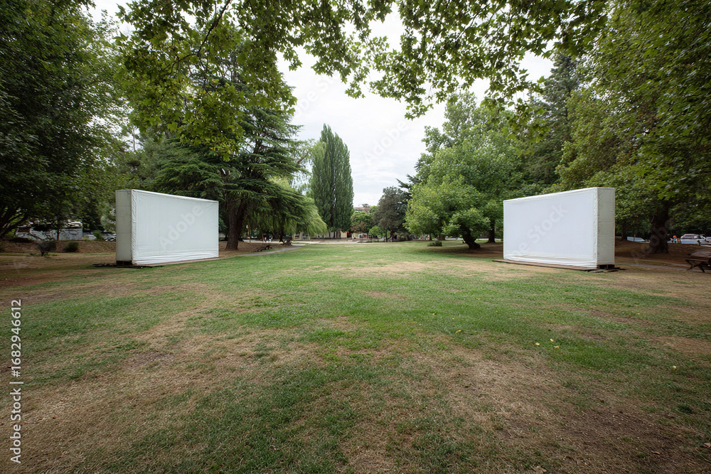 Fototapeta premium Two large white rectangles stand in a green field with various trees, suggesting an outdoor art installation or display area.