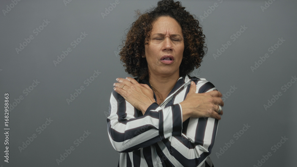 Naklejka premium Middle aged hispanic woman arms crossed hugging chest in gray studio wearing black and white striped blouse and rings, tense expression; cold discomfort.