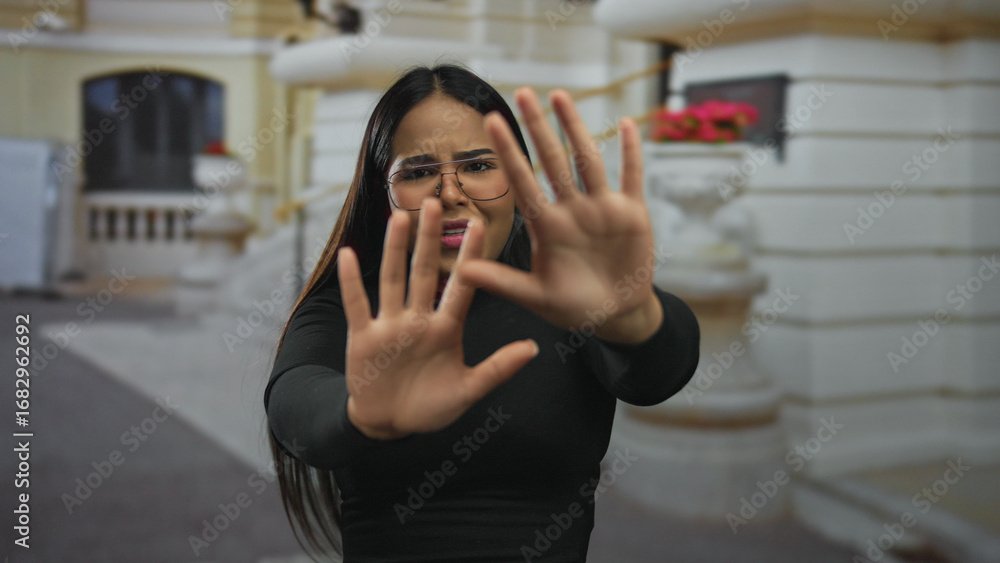 Fototapeta premium Woman with glasses gesturing stop in an urban outdoor setting showcasing expression with a blurred historic building background, emphasizing emotion and street environment.