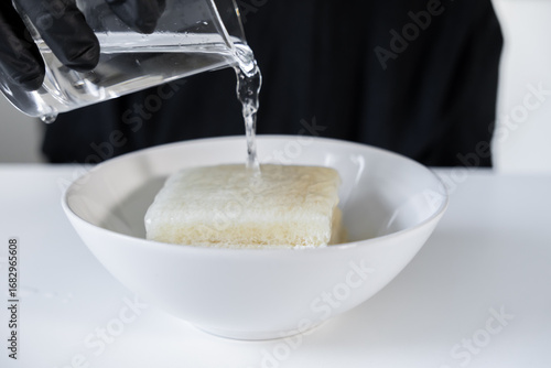 Photos Gloved hand pouring water over a block of bread soaking in a white bowl, essenti