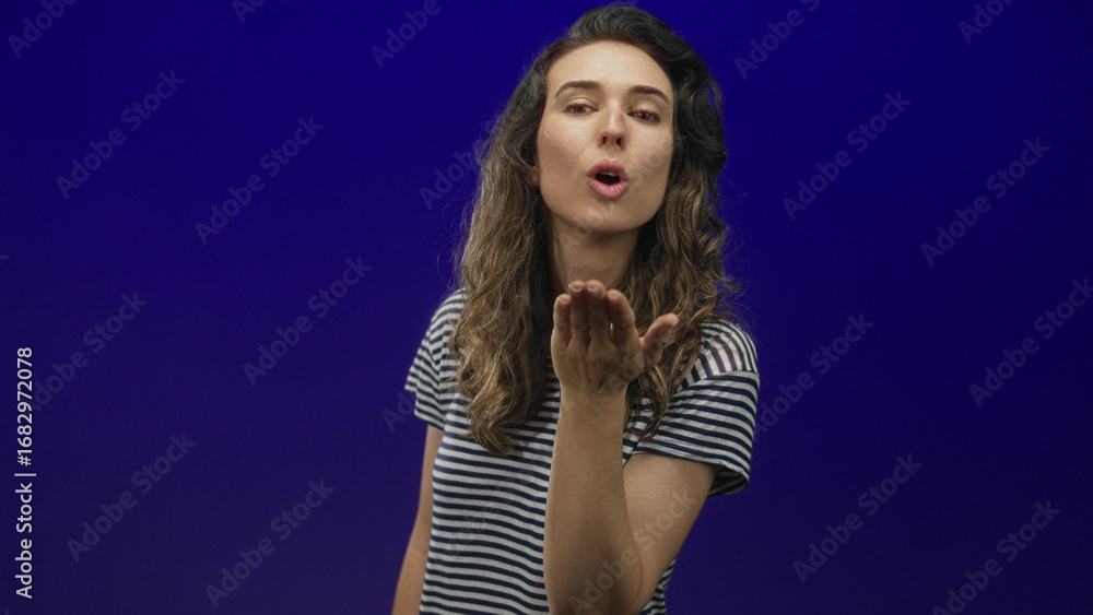 Naklejka premium Woman blows kiss with hand against blue studio wall while smiling softly with eyes closed; affection love.