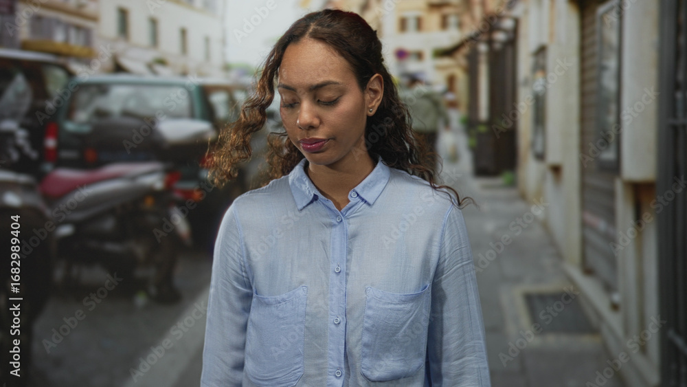 Fototapeta premium Woman with face visible and head bowed while looking down on a narrow urban street with parked scooters and cars; quiet melancholy.