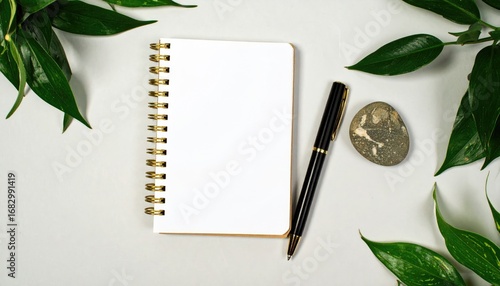 Minimalist desk setup with a blank spiral notebook, pen, and smooth stone surrounded by leafy green branches on a white surface