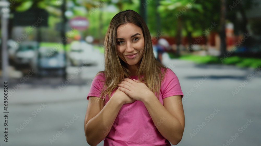 Fototapeta premium Woman in pink shirt stands on street with hands on chest, expressing emotion, surrounded by blurred background of trees, vehicles, and movement.