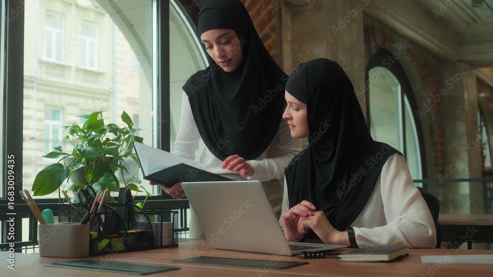 Two happy friendly muslim islamic businesswomen with laptop working teamwork colleagues girls female business women in hijab discuss project data together in office mentor helping intern with computer Two happy friendly muslim islamic businesswomen with laptop working teamwork colleagues girls female business women in hijab discuss project data together in office mentor helping intern with computer