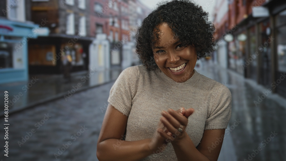 Fototapeta premium Young african american woman clapping hands and smiling broadly on a rain slicked city street lined with brick shops; celebration.